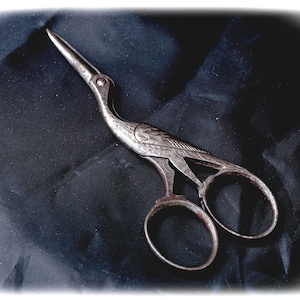 Può includere: Un paio di forbici in metallo color argento a forma di gru. Le lame formano il becco dell'uccello, mentre le impugnature sono progettate come la coda e il corpo. La superficie metallica presenta dettagli di piume incisi lungo il collo e le ali.
