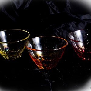 Puede incluir: Tres cuencos de vidrio transparente con bordes de colores, cada uno con un tono diferente: amarillo, naranja y rojo. Los cuencos tienen un diseño facetado y están colocados sobre un fondo oscuro, creando un contraste dramático. Los cuencos son probablemente para servir o exhibir.