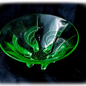Puede incluir: Un cuenco de cristal verde vibrante con un patrón de espiral en el centro de la base y líneas grabadas que irradian hacia el borde. El cuenco se apoya sobre tres pequeños pies de cristal integrados y muestra un tono verde brillante y translúcido.