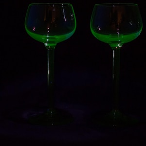 Puede incluir: Dos copas de tallo a juego con un tono verde neón vibrante y brillante. Cada copa tiene un cuenco redondeado, un tallo delgado y una base circular. Las copas están iluminadas sobre un fondo oscuro, resaltando su color verde brillante.