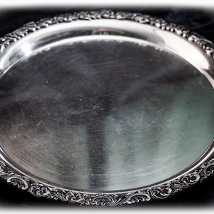 Puede incluir: Una bandeja de servicio redonda de plata con una superficie central pulida y reflectante, y un borde ancho y ornamentado con intrincados patrones de volutas y hojas en relieve. El texto "Torch & Talon" es visible abajo a la derecha.