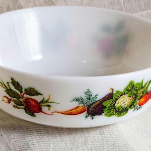 May include: White, oval-shaped serving bowl with two handles. The bowl features a colorful vegetable design around the exterior, including carrots, radishes, eggplants, and cauliflower. The bowl is likely made of ceramic or glass.
