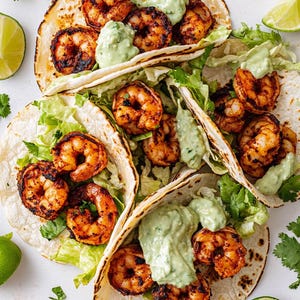 May include: Three shrimp tacos, viewed from above, on a white surface. Each taco contains grilled prawns, shredded lettuce, and a creamy green sauce, all within a warm tortilla. Lime wedges and coriander sprigs are scattered around.