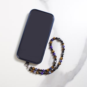 May include: A dark blue smartphone in a matching case with a beaded phone strap. The strap features a mix of brown, black, purple, and gray beads. The phone and strap are on a white marble surface.