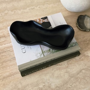 Peut inclure: Un plateau en céramique noir mat avec une forme organique irrégulière aux bords ondulés. Le plateau présente un bassin profond et lisse, posé sur deux livres à couverture rigide empilés intitulés Curate et Cocoon, avec une finition sombre.
