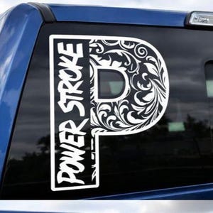 Puede incluir: Calcomanía de vinilo blanco con una gran letra "P" con un diseño floral ornamentado en el lado derecho. El lado izquierdo de la "P" tiene las palabras "POWER STROKE" en letras en negrita y estilizadas. La calcomanía está adherida a una ventana azul de un vehículo.