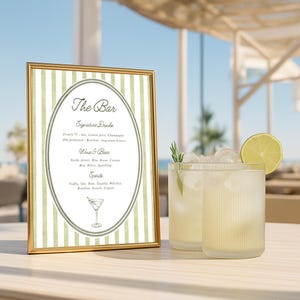 May include: A gold-framed sign with green and white stripes, displaying "The Bar" and drink options. Two glasses of cocktails with ice, lime, and rosemary sit on a wooden table, with a blurred ocean background.