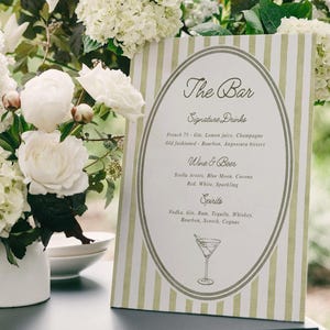 May include: A bar menu with a green and white striped design. The menu lists signature drinks, wine & beer, and spirits. White flowers and greenery surround the menu.