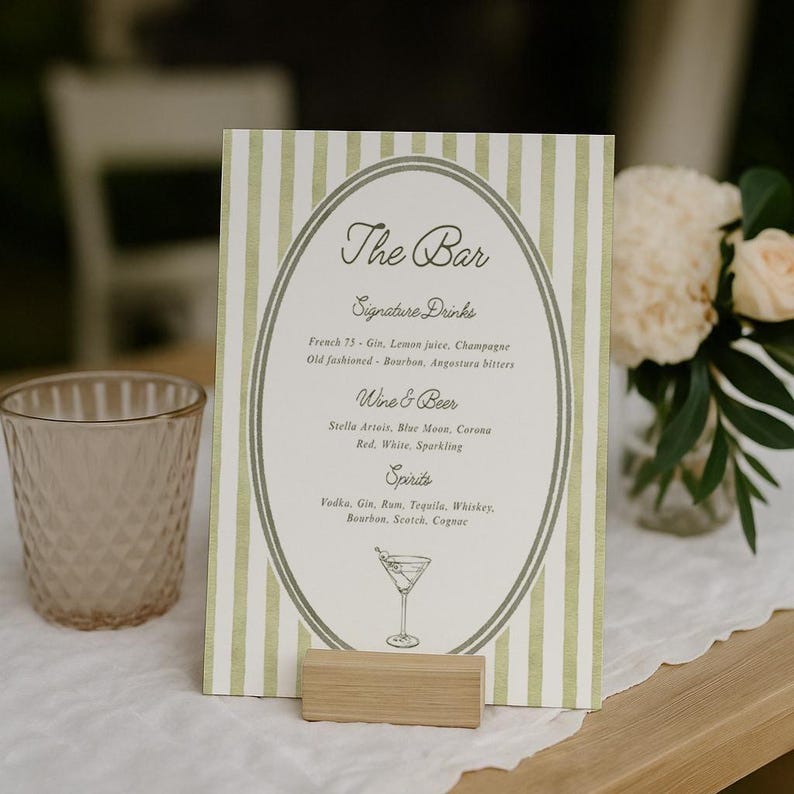 May include: A drinks menu with green and white striped background, listing cocktails and drinks. The sign is held up by a small wooden block. A clear glass and flowers are in the background. The menu includes 'Signature Drinks' and 'Spirits'.