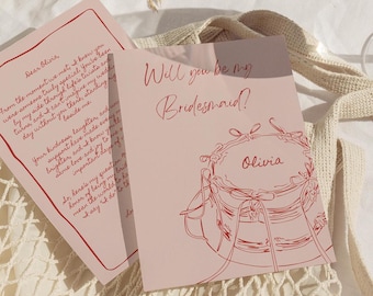 May include: A pink card with red text that says "Will you be my Bridesmaid?" with a drawing of a cake with a red ribbon and the name "Olivia" on it.