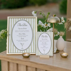 May include: A bar sign with a gold frame and green and white striped background. The sign reads "The Bar" with a list of signature drinks, wine, beer, and spirits. A smaller matching sign is displayed next to it. A gold lamp and candles are also on the table.