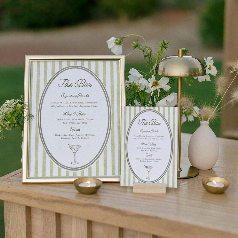May include: A bar sign with a gold frame and green and white striped background. The sign reads "The Bar" with a list of signature drinks, wine, beer, and spirits. A smaller matching sign is displayed next to it. A gold lamp and candles are also on the table.