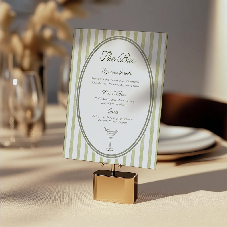 May include: A drinks menu card with a green and white striped design, held in a gold-coloured metal stand. The menu lists signature drinks, wine & beer, and spirits. The text includes "The Bar" and a cocktail illustration.
