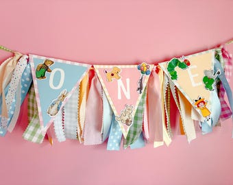 May include: A decorative fabric bunting banner featuring three triangular pennants spelling the word ONE. The pennants are decorated with various children's book character illustrations and are accented with hanging ribbons in pastel colors.