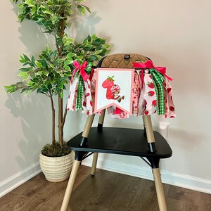 May include: A high chair skirt with a strawberry theme. The skirt features pink fabric with strawberry patterns, green gingham, and a banner with a strawberry character. The skirt is decorated with pink and green ribbons.