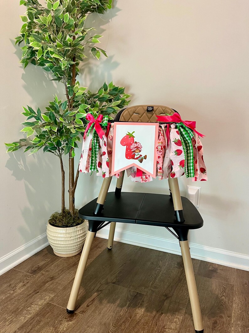 May include: A high chair skirt with a strawberry theme. The skirt features pink fabric with strawberry patterns, green gingham, and a banner with a strawberry character. The skirt is decorated with pink and green ribbons.