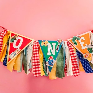 May include: A colorful banner spelling "ONE" with triangle flags and decorative ribbons. The flags are red, green, and orange, with the letters in white. The banner is decorated with illustrations of storybook characters and red and white checkered ribbons.