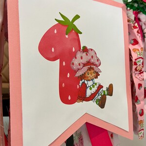 May include: A pink and white banner featuring a large red number one shaped like a strawberry. A cartoon character wearing a strawberry hat and dress is seated next to the number. The banner is trimmed in pink and has fabric accents.