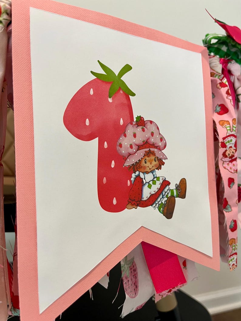 May include: A pink and white banner featuring a large red number one shaped like a strawberry. A cartoon character wearing a strawberry hat and dress is seated next to the number. The banner is trimmed in pink and has fabric accents.