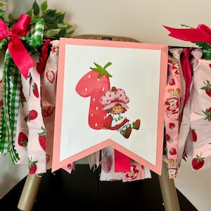 May include: A festive banner featuring a large number one shaped like a strawberry, with a cartoon character. The banner is decorated with pink, white, and green fabric strips, bows, and strawberry patterns, perfect for a first birthday celebration.