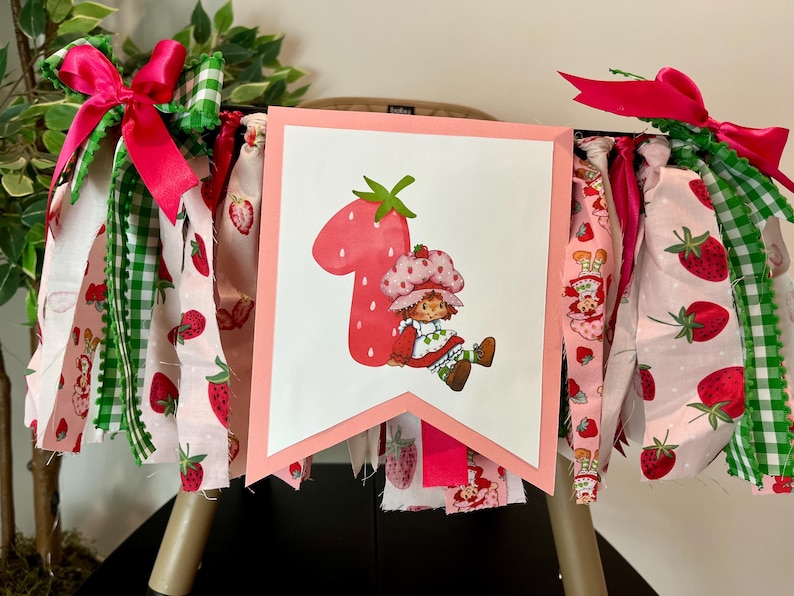 May include: A festive banner featuring a large number one shaped like a strawberry, with a cartoon character. The banner is decorated with pink, white, and green fabric strips, bows, and strawberry patterns, perfect for a first birthday celebration.