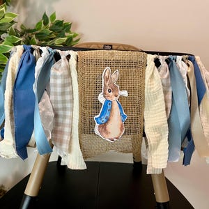 May include: A high chair skirt with a Peter Rabbit theme. The skirt has a burlap centre with a Peter Rabbit graphic, surrounded by blue, cream, and plaid fabric strips. The high chair legs are tan and black.