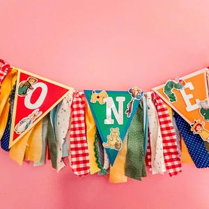 May include: A decorative fabric banner spelling the word ONE with three triangular pennants. The red, green, and orange pennants feature classic storybook character illustrations and silver letters, accented by hanging strips of colorful fabric.