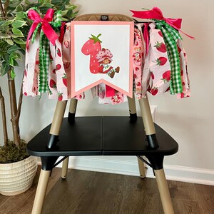May include: A high chair decorated with a banner featuring a strawberry and a cartoon character. The banner is framed in pink and adorned with pink and green ribbons. The chair is black with tan legs, and the banner is decorated with pink fabric with strawberries.
