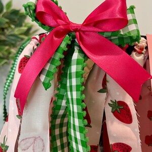 May include: A close-up of a decorative bow made of bright pink satin ribbon, atop green and white gingham ribbon with green pom-pom trim. The background features fabric with strawberry patterns.