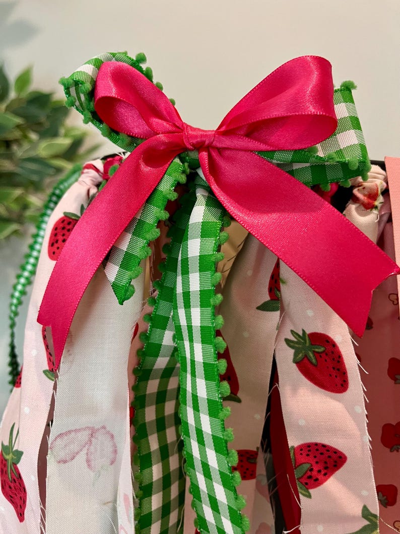 May include: A close-up of a decorative bow made of bright pink satin ribbon, atop green and white gingham ribbon with green pom-pom trim. The background features fabric with strawberry patterns.