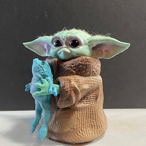 May include: A green and blue figurine of a character from the Star Wars franchise, holding a blue frog in its hands. The character is wearing a brown robe.