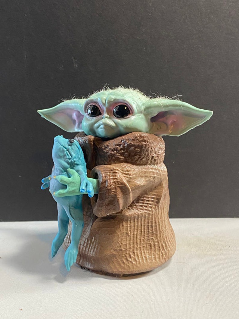 May include: A green and blue figurine of a character from the Star Wars franchise, holding a blue frog in its hands. The character is wearing a brown robe.