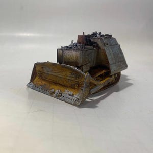 May include: A miniature, weathered bulldozer model with a rusted yellow blade and a gray body. The model has a detailed design, including tracks and a cab filled with debris. The overall aesthetic suggests a post-apocalyptic or industrial theme.