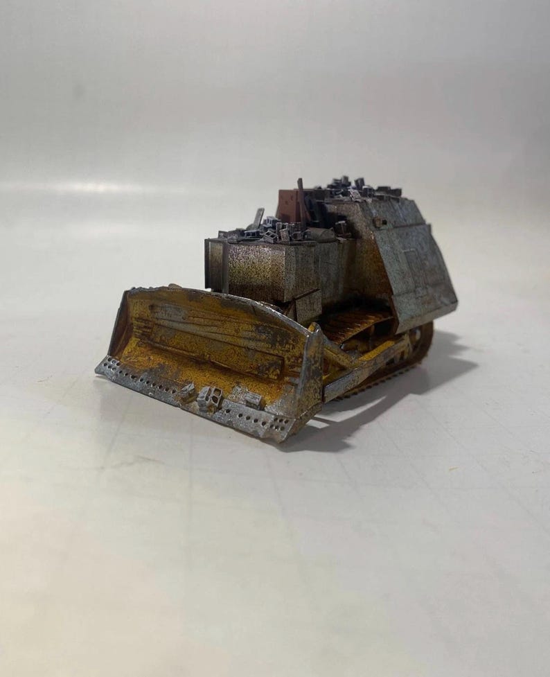 May include: A miniature, weathered bulldozer model with a rusted yellow blade and a gray body. The model has a detailed design, including tracks and a cab filled with debris. The overall aesthetic suggests a post-apocalyptic or industrial theme.