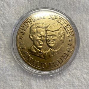 以下が含まれることがあります： 透明なプラスチックホルダーに入った金色のコイン。コインには2人の男性の肖像画と「JEFFREY EPSTEIN」と「DONALD TRUMP」の文字が円形に配置されています。コインは光沢のある反射面を持っています。