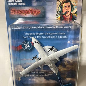May include: A white toy airplane with black propellers and a blue stand is displayed in a clear plastic package. The package features text that reads "Sky King Richard Russel" and "SavageRigs." The package also includes a picture of a man wearing a headset.