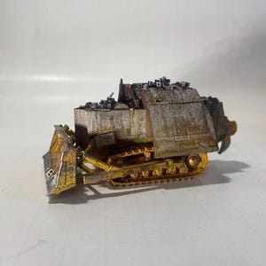 May include: A miniature, weathered bulldozer model. The model features a yellow blade and tracks, with a rusted, metallic body. The top of the bulldozer is covered with small, dark-colored details, possibly representing debris or equipment.