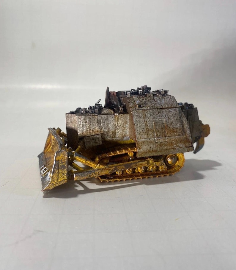 May include: A miniature, weathered bulldozer model. The model features a yellow blade and tracks, with a rusted, metallic body. The top of the bulldozer is covered with small, dark-colored details, possibly representing debris or equipment.