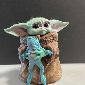 May include: A small green and blue figurine of a character from the Star Wars franchise, holding a blue frog figurine. The character has large eyes and is wearing a brown robe.