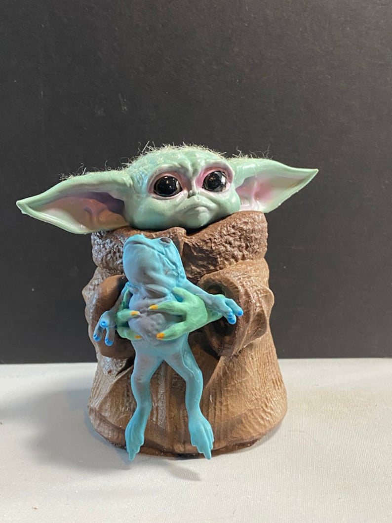 May include: A small green and blue figurine of a character from the Star Wars franchise, holding a blue frog figurine. The character has large eyes and is wearing a brown robe.