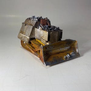 May include: A miniature bulldozer model with a weathered, rusty appearance. The blade and tracks are yellow and brown, with a silver and brown body. The top of the bulldozer is covered with small, gray rectangular shapes.