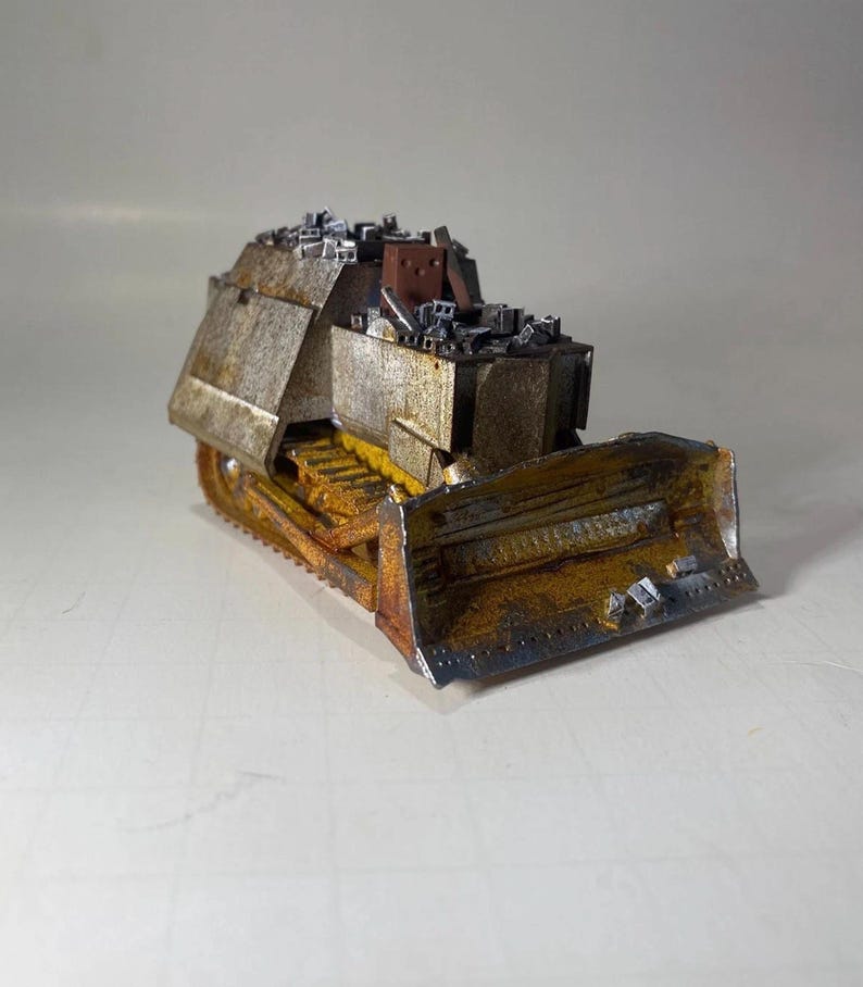 May include: A miniature bulldozer model with a weathered, rusty appearance. The blade and tracks are yellow and brown, with a silver and brown body. The top of the bulldozer is covered with small, gray rectangular shapes.