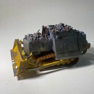May include: A miniature model of a bulldozer, featuring a yellow blade and tracks, with a dark gray body. The model has a detailed design, including a small structure on top, and is set against a plain background.