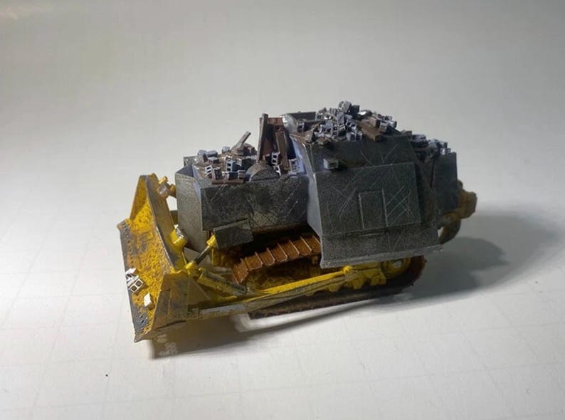 May include: A miniature model of a bulldozer, featuring a yellow blade and tracks, with a dark gray body. The model has a detailed design, including a small structure on top, and is set against a plain background.