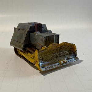 May include: A miniature model of a bulldozer, featuring a yellow blade and tracks, with a gray cab and body. The model has a weathered appearance, suggesting it is a vintage or antique piece. The bulldozer is set against a neutral background.