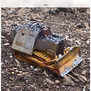 May include: A detailed model of a bulldozer, featuring a yellow blade and tracks, with a weathered, metallic finish. The model is set against a backdrop of wood chips and small debris, showcasing its construction and design.