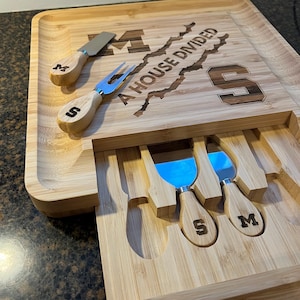 May include: A bamboo cheese board set with a slide-out drawer. The board is engraved with the text "A HOUSE DIVIDED" and the letters "M" and "S". The set includes three cheese knives and a cheese fork, each with a wooden handle.