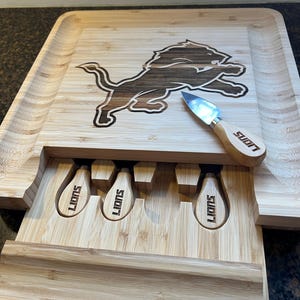 May include: A wooden cutting board with a lion logo and a drawer that holds four cheese knives. The knives have the word "Lions" engraved on them.