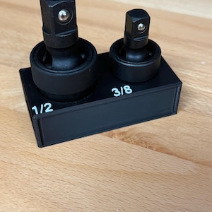 May include: A black plastic storage holder for two universal joint socket adapters. The holder features white printed text labels reading 1/2 and 3/8, corresponding to the two black metal swivel adapters inserted into the top of the base.