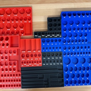 May include: A collection of red, black, and blue plastic organizers for tools. The organizers have various sized circular holes and rectangular slots, with measurements printed on them. The tools organizers are arranged on a light wood surface.